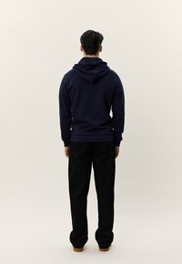 Homem de costas para a câmera, vestindo um capuz azul-marinho, calças pretas e sapatos castanhos contra um fundo claro e simples.