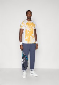 Športový outfit pozostávajúci z krátkeho bieleho trička s žltými geometrickými vzormi, námorníckych nohavíc a bielych tenisiek, držiaci tenisovú raketu.