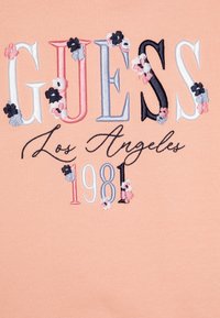 Perzikkleurige katoenstof met geborduurde tekst "GUESS Los Angeles 1981" in meerkleurige letters en bloemenaccenten.