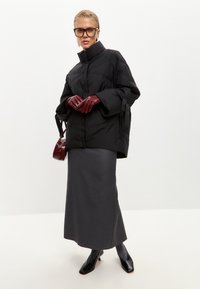 Giacca a vento nera con collo alto, abbinata a una gonna grigia lunga fino a terra. Gli accessori includono guanti bordeaux e una piccola borsa. Scarpe nere.