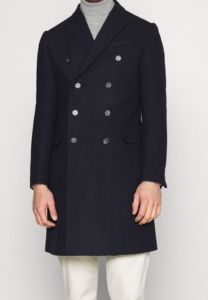 Homme portant un manteau en laine noir à double boutonnage sur un col roulé gris et un pantalon blanc, debout devant un fond uni.