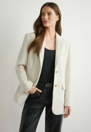 Kobieta z długimi brązowymi włosami, ubrana w biały żakiet, czarną bluzkę, czarne spodnie i pasek z podwójnymi kółkami, stojąca z jedną ręką w kieszeni.