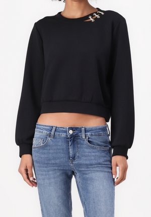 Kvinde iført sort cropped sweatshirt med lange ærmer og sølvkædedetalje ved halsudskæringen samt blå jeans med normal talje.