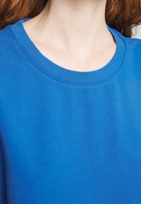 Sininen lyhythihainen t-paita, jossa on pyöreä kaula-aukko, sileä rakenne ja tasainen kangas. Ompeleet kaula-aukossa lisäävät rakennetta.