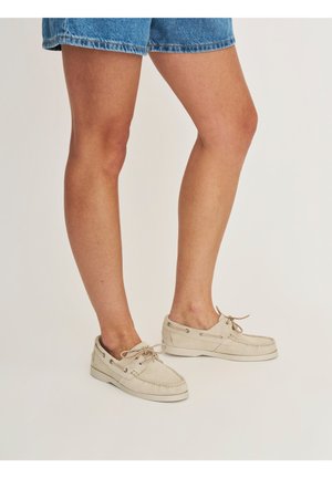 Benen met beige suède bootschoenen met veters, gecombineerd met blauwe denim shorts, op een eenvoudige lichte achtergrond.