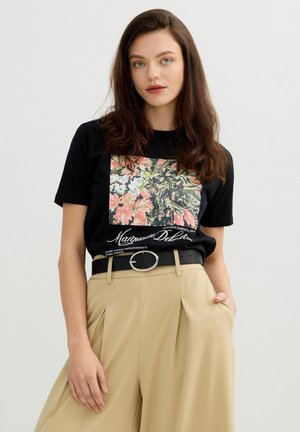 Joven mujer con cabello largo y castaño lleva una camiseta negra con estampado floral metida en pantalones beige de cintura alta con cinturón negro, de pie con una mano en el bolsillo.