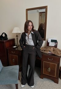 Femme aux cheveux bruns portant un costume sombre et une chemise claire, debout les mains dans les poches près d'un bureau en bois et d'un miroir mural dans une pièce.