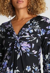 Robe noire à fleurs avec des manches longues. Présente des motifs floraux violets et bleus, un design frontal torsadé et un décolleté en V.