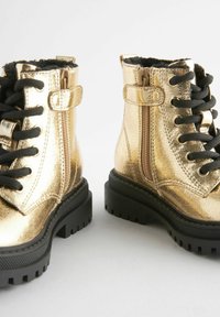 Goldene metallische Knöchelstiefel mit schwarzen Schnüren, Reißverschluss und strukturiertem Finish. Sie verfügen über eine klobige schwarze Gummisohle mit Profil.