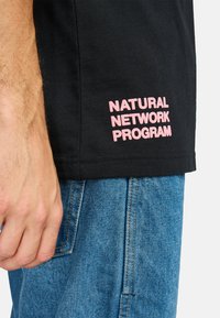 Black cotton t-shirt with pink embroidered text "NATURAL NETWORK PROGRAM" on the side. Casual fit, worn with light blue denim pants.