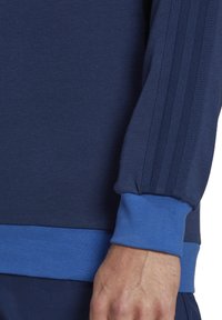 Marineblauwe pullover met geribbelde textuur, contrasterende lichtblauwe manchetten en zoom, met drie verticale strepen op de mouw.