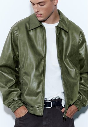 Homme portant une veste en cuir vert olive ouverte sur un t-shirt blanc, les mains dans les poches d’un pantalon foncé.