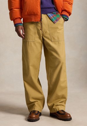 Homme portant un pantalon large moutarde, des chaussures de randonnée marron, une doudoune orange, un pull violet et une chemise à carreaux avec une main dans la poche.