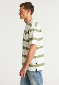 Balta marškinėliai su alyvų žalios horizontaliomis juostelėmis, apvaliu kaklu, krūtinės kišene ir šoninėmis plyšimais apačioje.