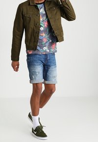 Chaqueta de mezclilla verde sobre una camiseta floral, combinada con pantalones cortos azules desgastados. Calcetines blancos y zapatillas verdes completan el conjunto.