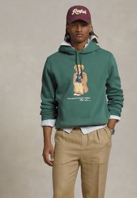 Groene hoodie met een berenafbeelding en de tekst "Polo Bear by Ralph Lauren". Gecombineerd met tan-kleurige geruite broek. Witte geknoopte shirt eronder.