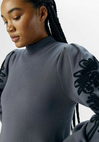 Pull en tricot gris avec un col montant, doté de manches bouffantes et d'une broderie florale noire sur les épaules. Texture douce et design ajusté.