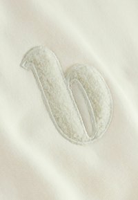 Het nummer "10" geborduurd met witte, donzige textuur draad op een zachte, licht romige stof achtergrond.