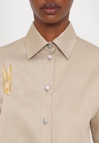 Beige button-up shirt with a pointed collar, featuring metallic buttons and a yellow and gray safety pin embroidery on the chest.