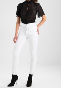 Blusa negra transparente con mangas cortas y volantes, y detalle de botones, combinada con jeans skinny blancos de tiro alto y botines negros de tacón.
