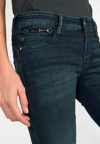 Jean en denim de couleur foncée avec une coupe ajustée. Les caractéristiques incluent une poche avec un accent en chaîne décorative et un léger effet de délavage sur le tissu.