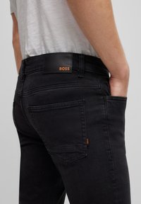 Persona que lleva unos pantalones BOSS negros con el bolsillo trasero visible y la mano derecha en el bolsillo delantero, combinados con una camiseta blanca.