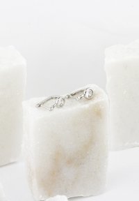 Silverörhängen med en rund, klar ädelsten, som har en låsdesign med hävarm. Stenen är inställd i en slät sarg, på en ljus bakgrund.