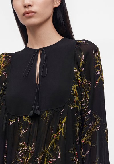 Femme portant une robe noire à fleurs avec des manches bouffantes en voile transparent et un large plastron noir uni noué au niveau du col avec des cordons à pompons.