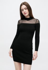 Černé žebrované pletené šaty s dlouhými rukávy, průhledným síťovaným výstřihem, ozdobené malými aplikacemi podél ramen.