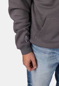 Felpa grigia con polsini a costine e tasca frontale, con un piccolo logo geometrico bianco sul polsino. Un paio di jeans in denim blu sono visibili.