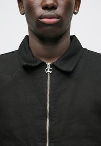 Chaqueta negra con cremallera, confeccionada en tejido texturizado; cuenta con un cuello con puntas y una cremallera plateada como acento. Vista en primer plano de la prenda superior.