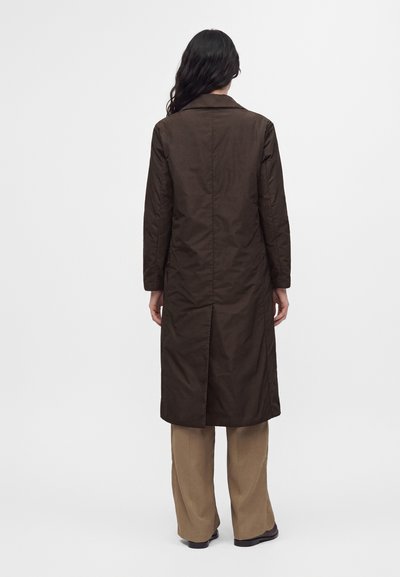 Femme aux longs cheveux foncés portant un manteau brun foncé jusqu'aux genoux, un pantalon beige et des chaussures noires, debout face à un fond blanc.