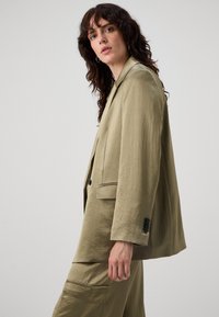 Blazer oversize khaki con finitura testurizzata, due tasche frontali e bottoni neri, indossato sopra pantaloni coordinati.