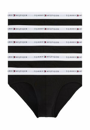 Uomo muscoloso in piedi a piedi nudi con slip neri con fascia bianca Tommy Hilfiger in vita, guarda di lato con una mano sul fianco.