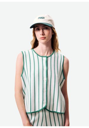 Femme portant un gilet blanc sans manches boutonné et un pantalon à rayures verticales vertes, ainsi qu'une casquette blanche à visière verte avec le texte "FRNCH".