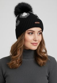 Black knitted beanie with white and gray heart patterns, featuring a fluffy black pom-pom on top and a brown leather logo patch.
