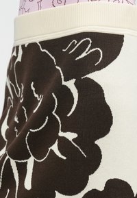 Detajl krila s črno-belim cvetličnim vzorcem in kremasto barvnim pasom, kombinirano s roza zgornjim delom z črnim linijskim dizajnom.