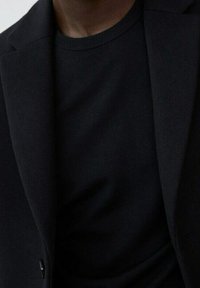 Schwarzer Blazer über einem schwarzen Rundhalsausschnitt-Shirt. Die Materialien wirken glatt mit subtiler Textur und ohne sichtbare Muster oder Hardware-Details.