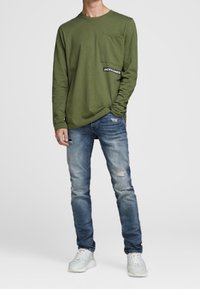 Man i en olivgrön långärmad skjorta med bröstficka och urvuxna blå jeans, står i vita sneakers på en enfärgad bakgrund.