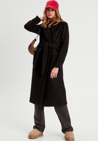 Black, textured long coat with a tie waist, worn over gray pants and tan slip-on shoes. A red cap and a leopard print bag add accents.