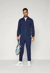 Man in navy tracksuit and white sneakers holding tennis racket and ball, standing against plain light background.