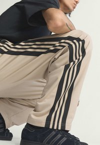 Béžové športové nohavice s textúrovaným materiálom, so tromi čiernymi pruhmi vertikálne po stranách. Vhodné s čiernymi teniskami.