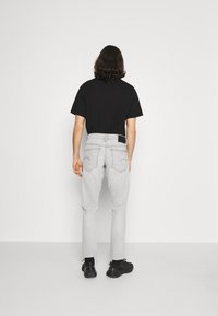 Hombre con el cabello hasta los hombros, vistiendo una camiseta negra, jeans gris claro y zapatillas negras, de pie frente a una pared blanca en blanco.