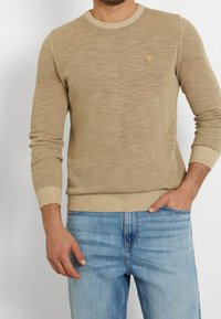 Uomo che indossa un maglione a maglia beige testurizzato e jeans di denim azzurri chiari, con una mano in tasca e l'altra rilassata al suo fianco.