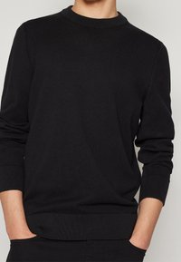 Černý svetr s kulatým výstřihem, který má textura pleteniny, žebrované manžety a lem. Design je jednoduchý a přiléhavý, bez viditelných log.