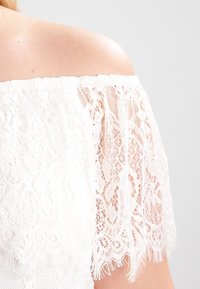 Haut blanc à épaules dénudées présentant des détails en dentelle complexe et une finition texturée. La dentelle possède des motifs floraux et des bords frangés.