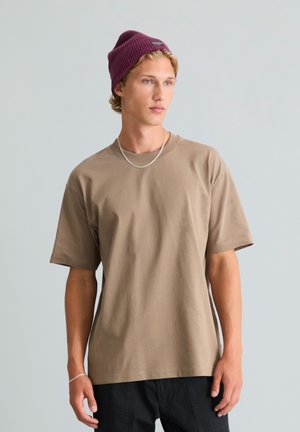 Jeune homme portant une chemise marron à manches courtes, un pantalon noir, un bonnet violet ainsi qu'un collier et un bracelet en chaîne argentés, debout devant un fond uni.