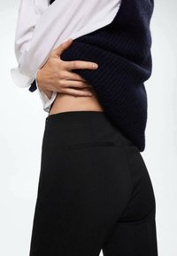 Black pants with a fitted design, featuring a back pocket and smooth texture. Paired with a dark, knitted top layered over a white shirt.