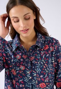 Donna con lunghi capelli castani che indossa una camicia a bottoni floreale blu navy, si tocca i capelli e guarda verso il basso con un sorriso dolce.