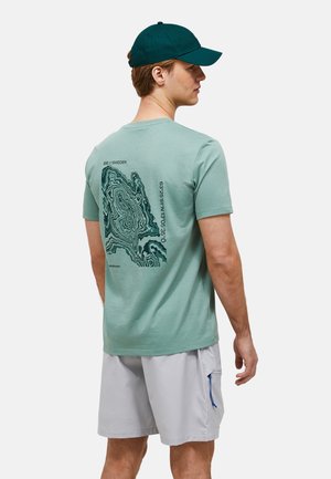 Ung mand iført en grøn kasket, grøn t-shirt med topografisk kort og koordinater printet på ryggen, og lysegrå shorts.